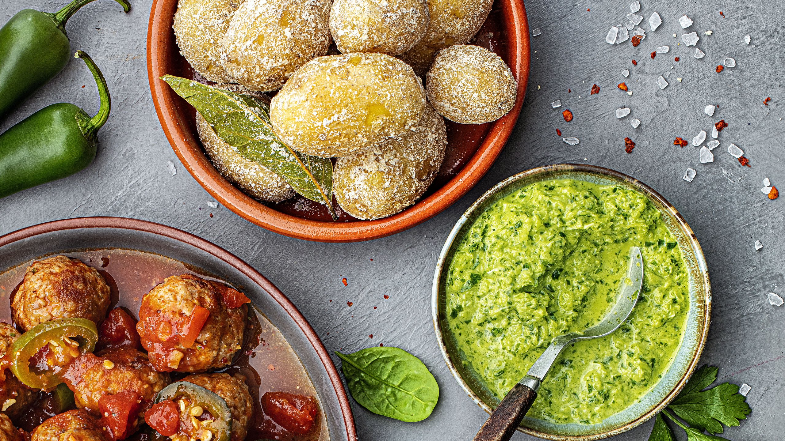 Links eine Schüssel mit Pesto, oben mittig eine Schüssel mit Kartoffeln, unten rechts Fleischbällchen mit Paprikasauce. Auf hellgrauem Untergrund mit Salzkörnern und Chillischoten