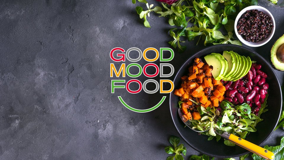 Rechts im Bild eine Feldsalat Bowl. Drumherum einige Salatblätter und Avocado, stehend auf einem dunklen Untergrund. Mittig das GOOD MOOD FOOD Logo.