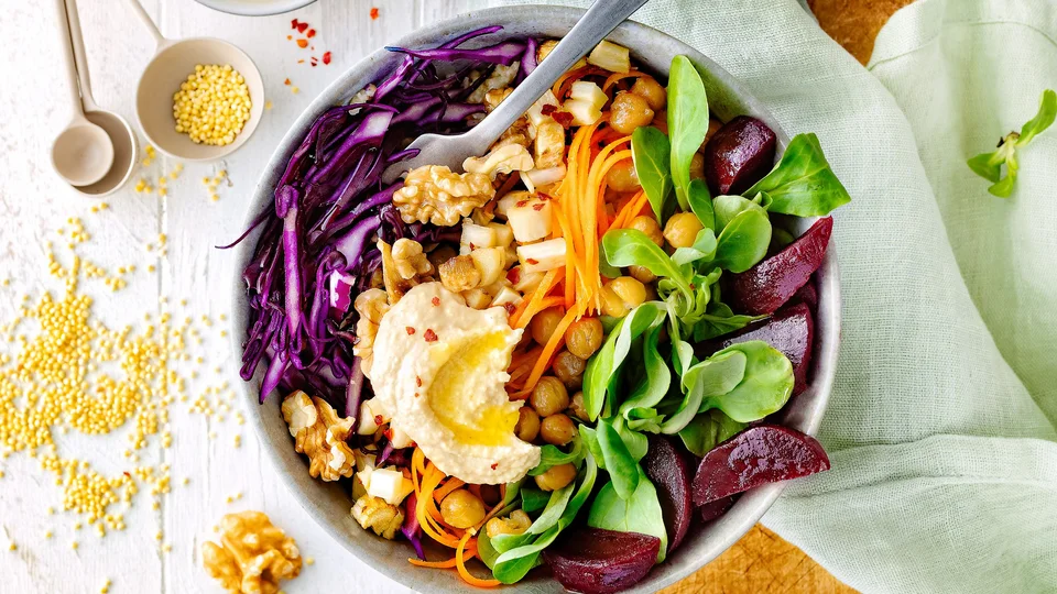 Eine Bowl aus Rotkohl, Kichererbsen, Feldsalat, Rote Beete und Walnüssen in einer weißen Schüssel. Links der Schüssel Quinoa und rechts ein hellgrünes Tuch. 