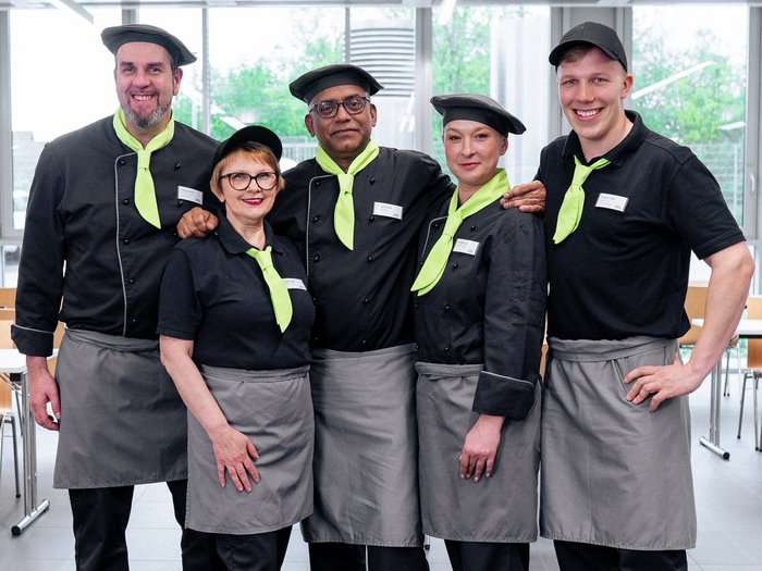 Ein Team aus fünf Köchen, welche in einer Kantine stehen. Sie tragen alle eine schwarze Kochjacke, eine schwarze Kochmütze und ein grünes Halstuch. zudem tragen sie eine graue Schürze 