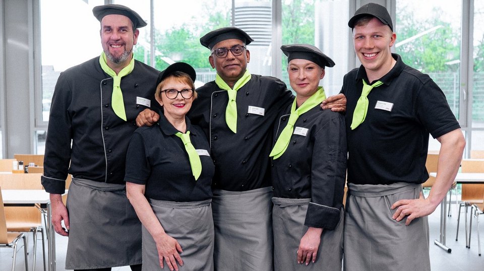 Ein Team aus fünf Köchen, welche in einer Kantine stehen. Sie tragen alle eine schwarze Kochjacke, eine schwarze Kochmütze und ein grünes Halstuch. zudem tragen sie eine graue Schürze 