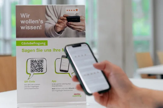 Eine Peron hält ihr Iphone, dahinter der QR-Code von easy pay, im Hintergrund weitere Stühle 