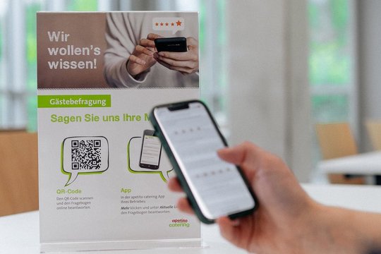 Eine Peron hält ihr Iphone, dahinter der QR-Code von easy pay, im Hintergrund weitere Stühle 